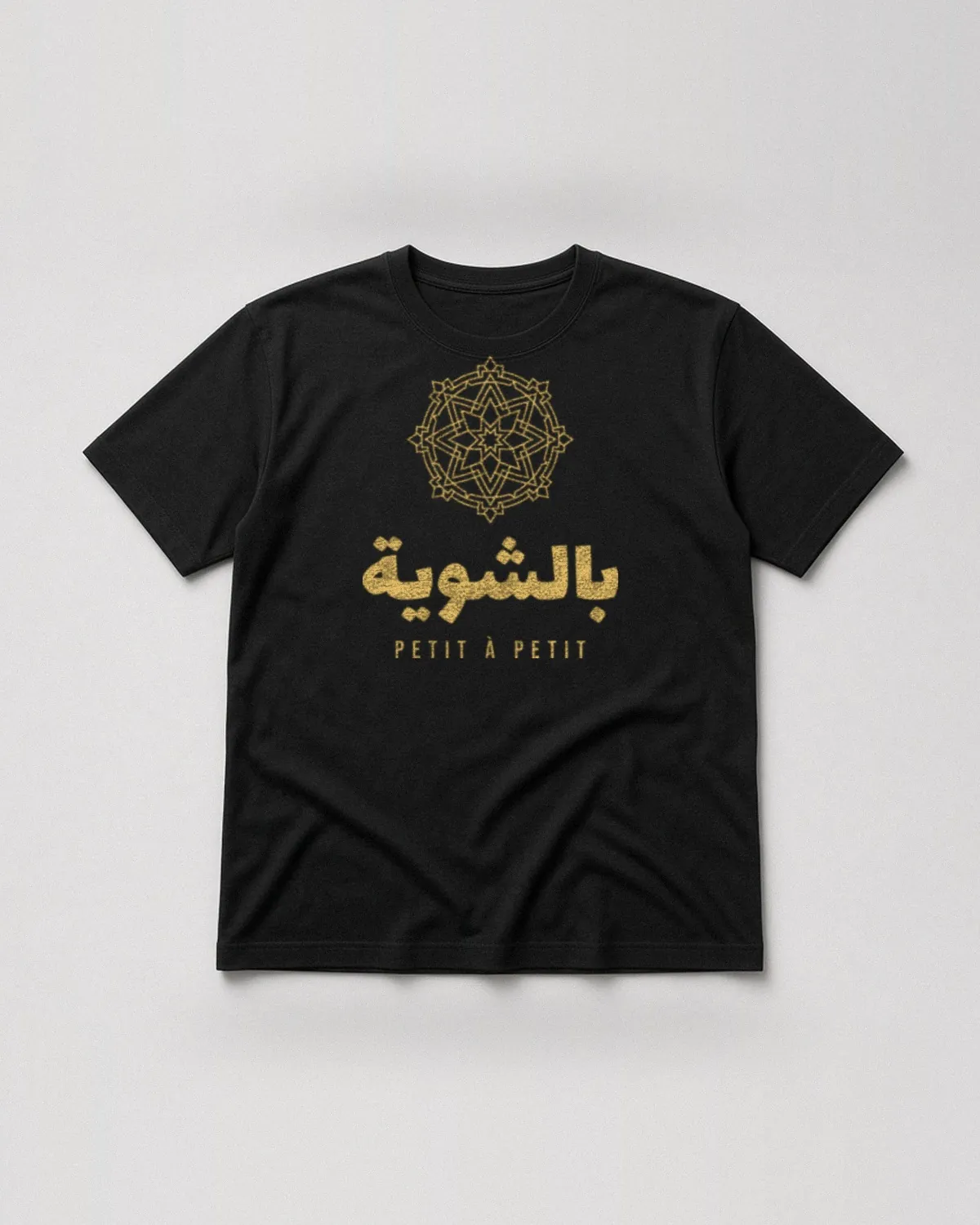 T-shirt noir avec le design « Bchwiya » : le mot arabe بالشوية sous une rosace géométrique, avec « Petit à petit » (vue de face, à plat)